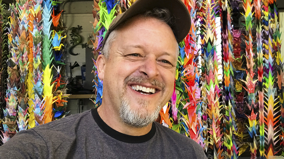 Rick Allred wearing a cap smiles with colourful origami cranes in the background.